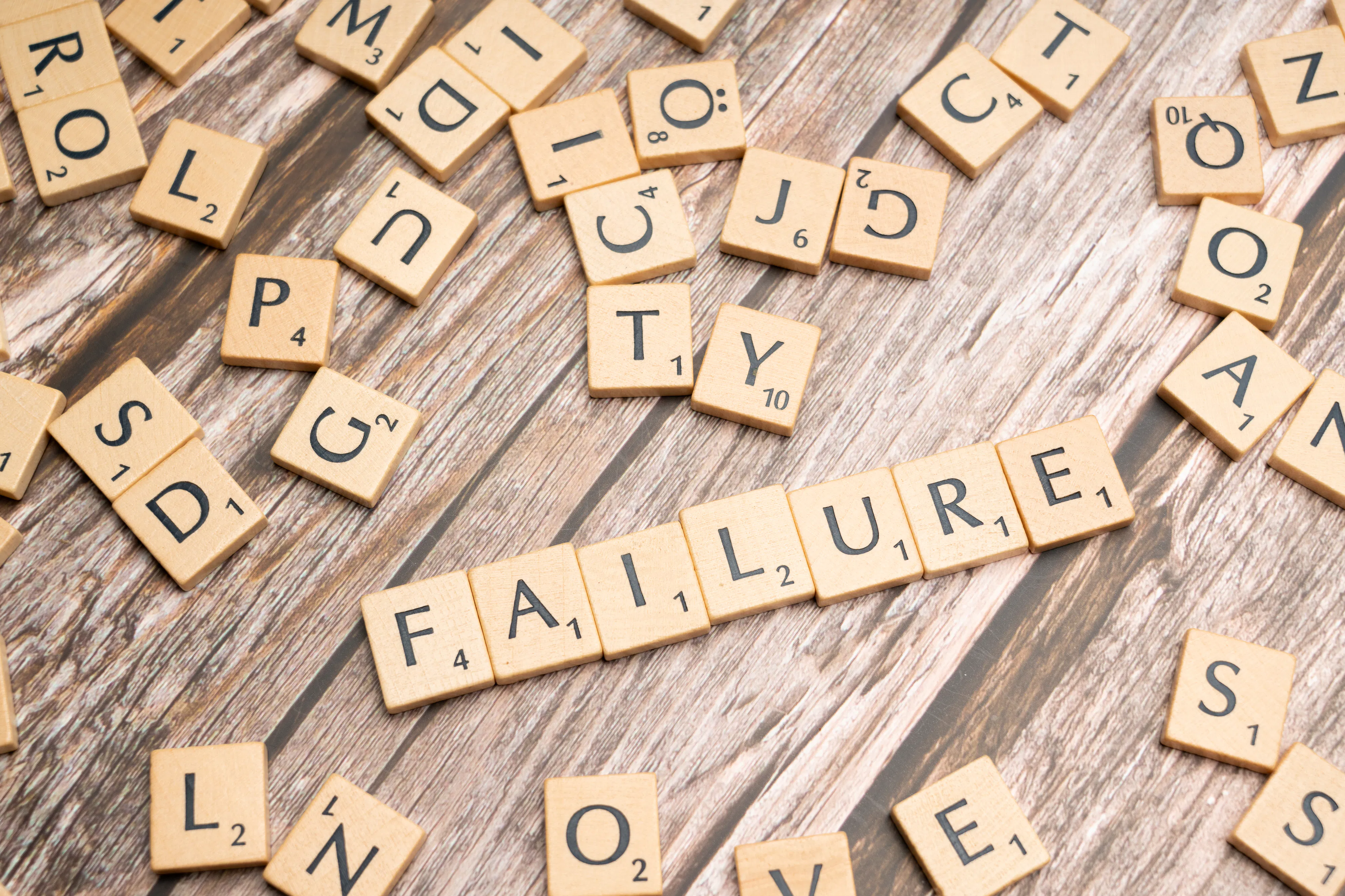 Scrabble tiles spelling out 'FAILURE' surrounded by other letter tiles on wood.