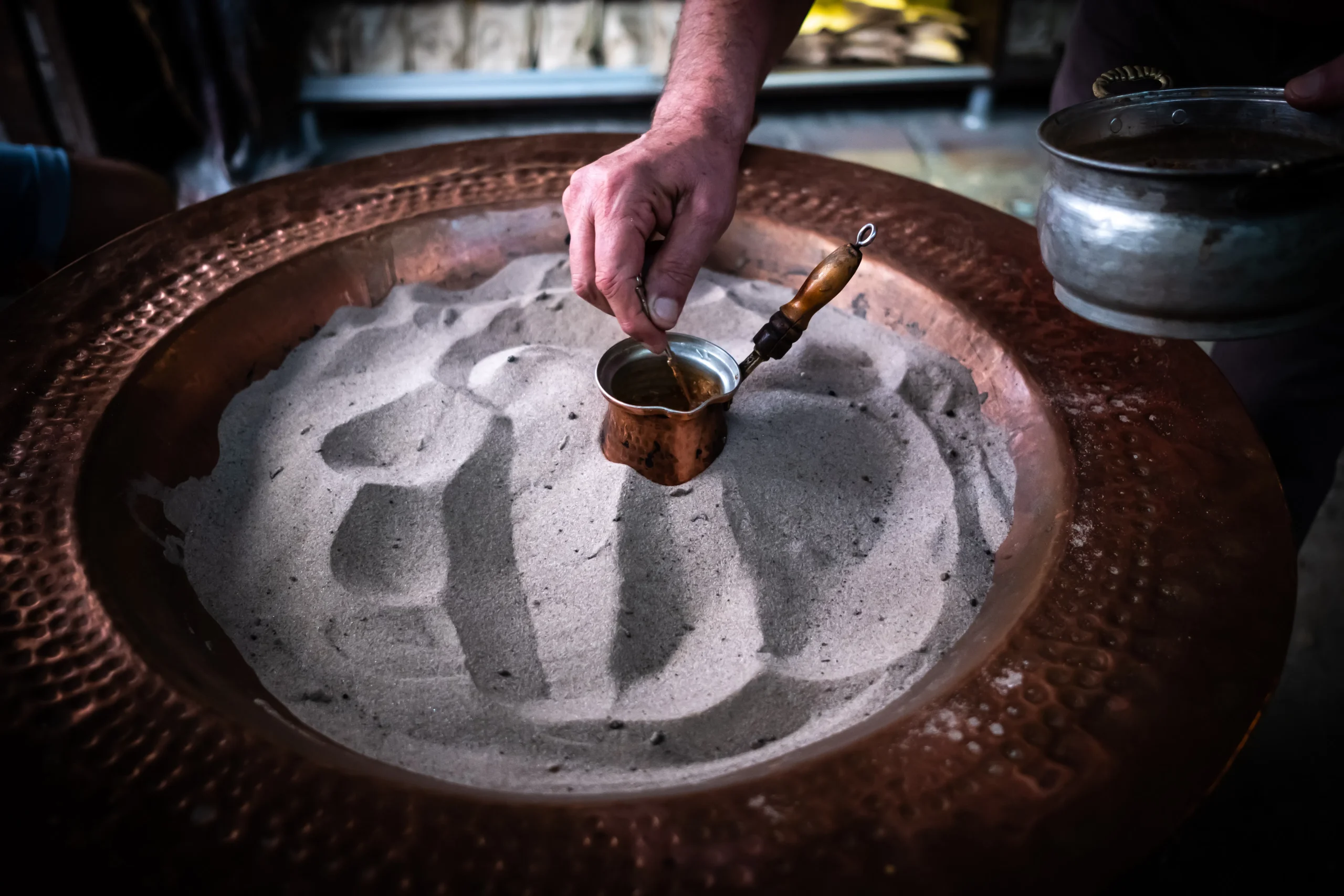 Close-up of hand preparing coffee in sand, using traditional pot.