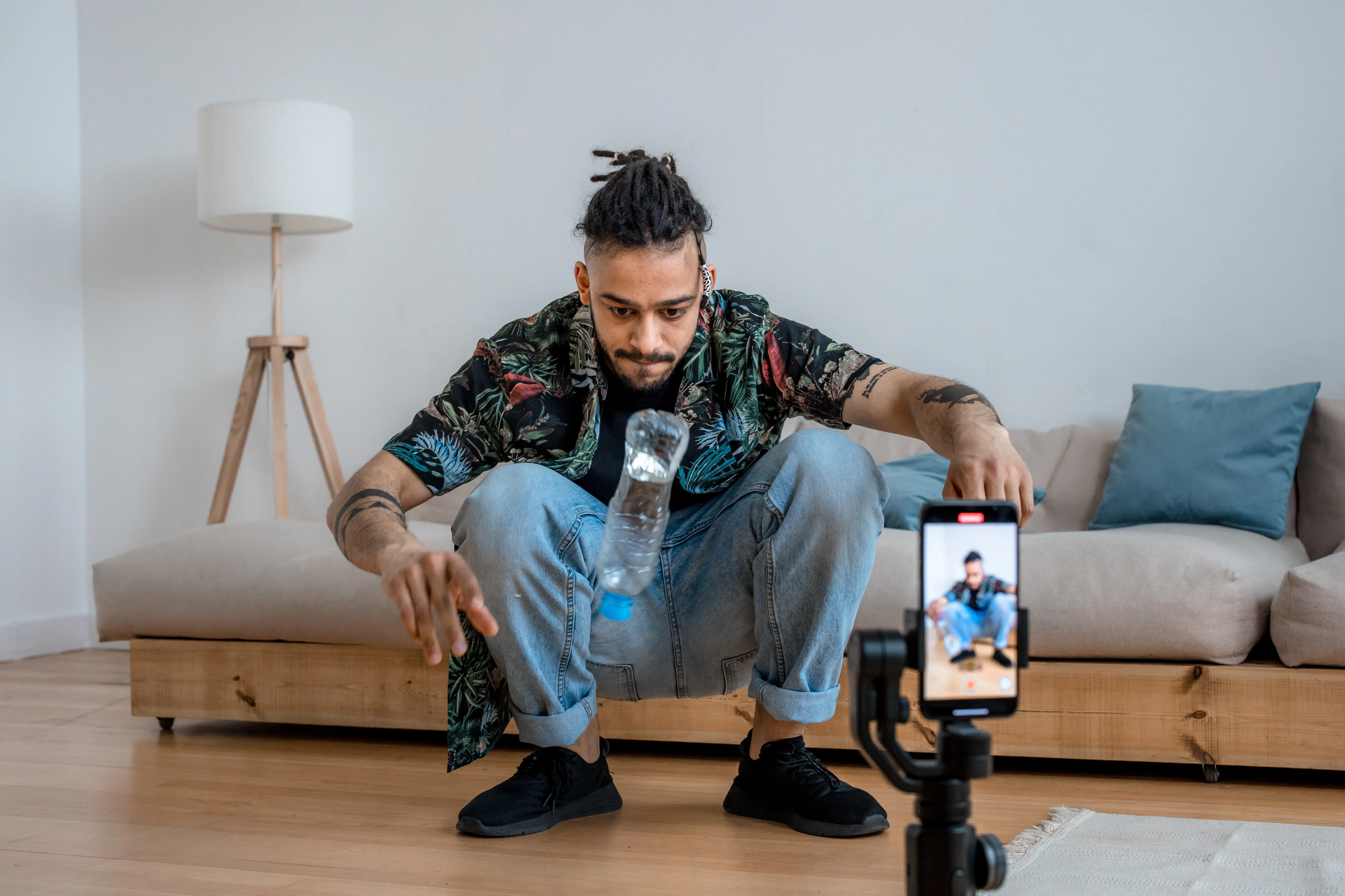 Man filming a water bottle flip challenge on a smartphone tripod indoors.