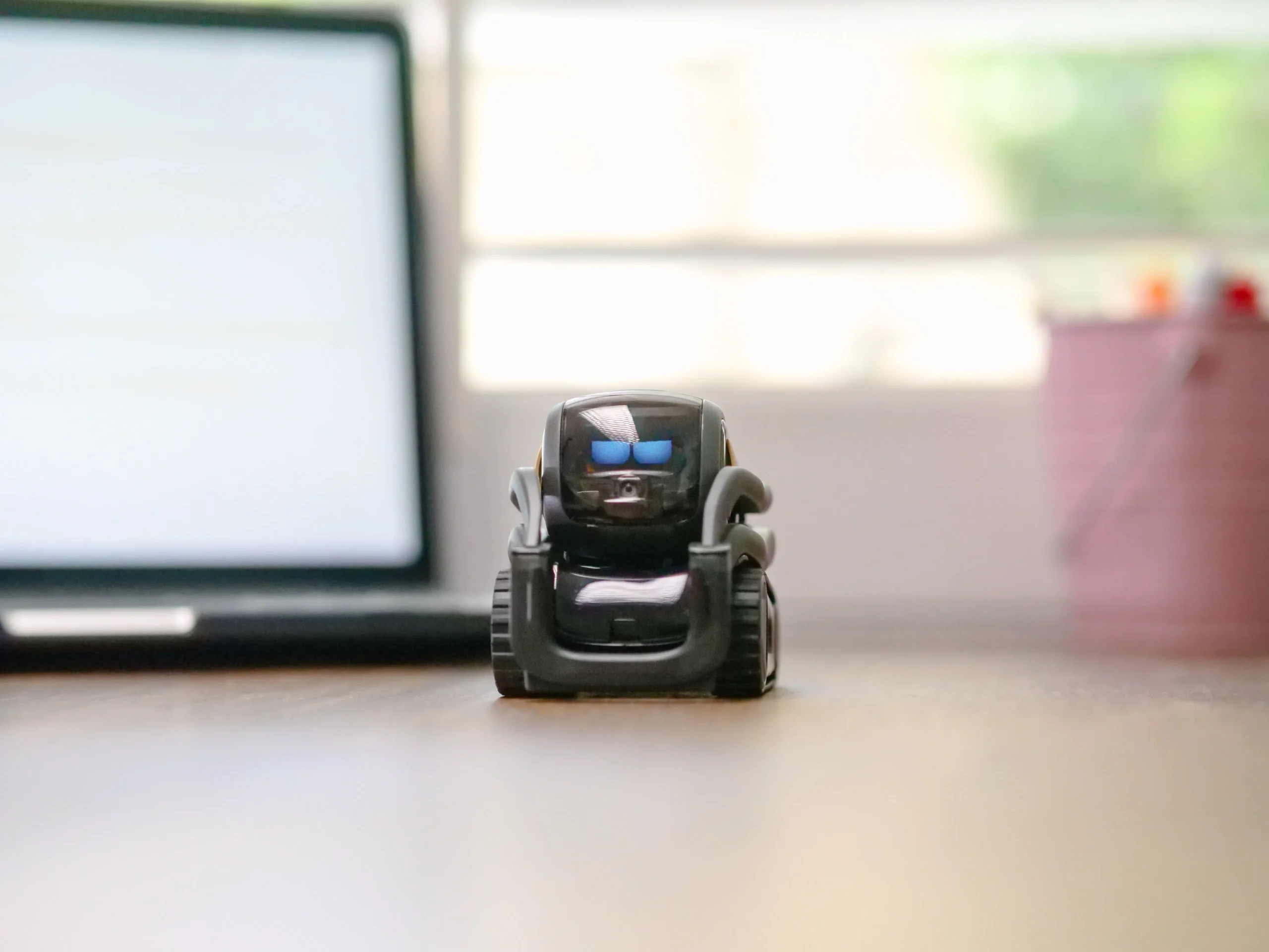 Close-up of a small AI robot on a desk with a laptop in the background.