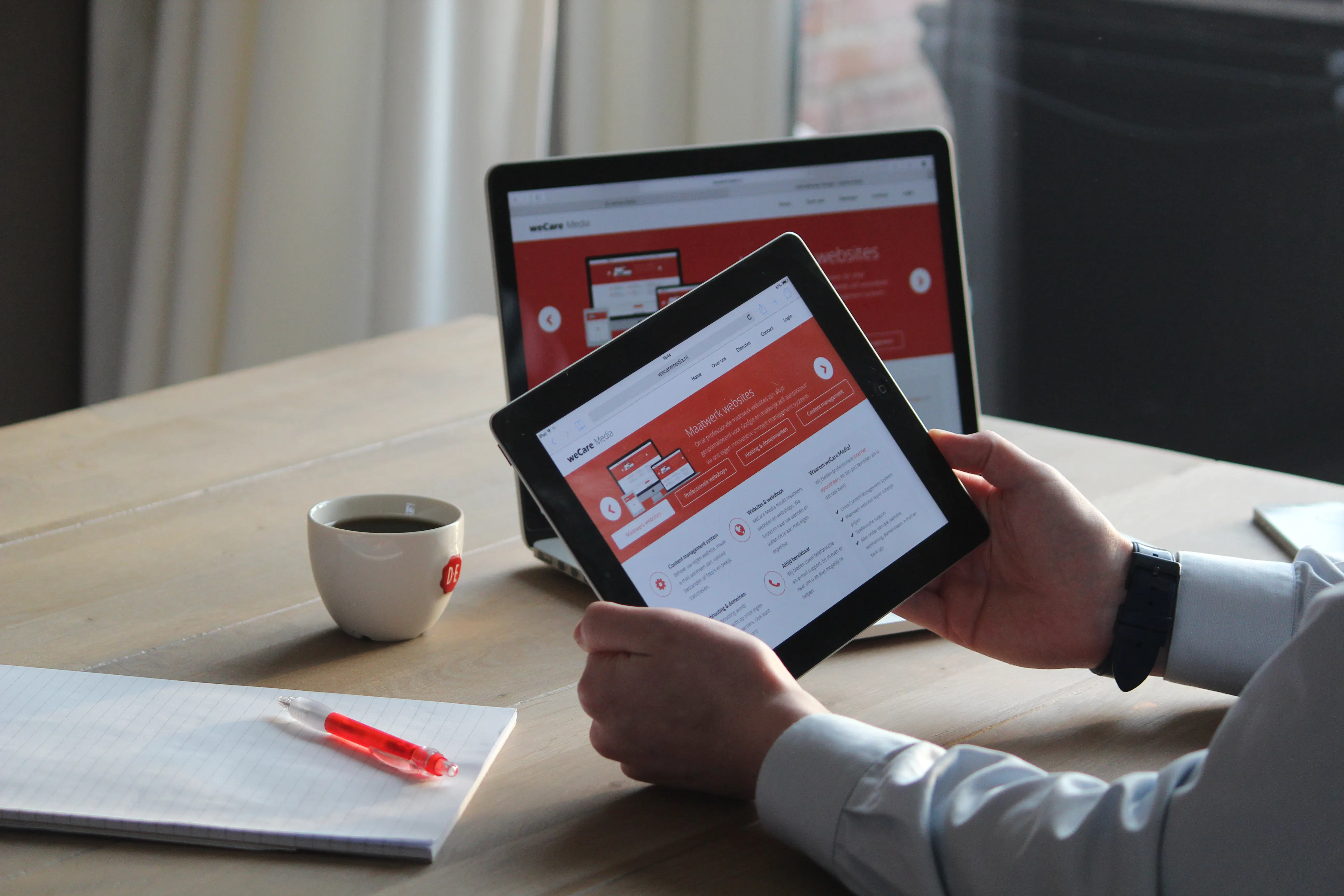 Person holding a tablet displaying a red and white website, with a laptop showing the same site in the background. A coffee cup, notebook, and red pen are on a wooden table, illustrating a marketing agency using an AI writing assistant for content.