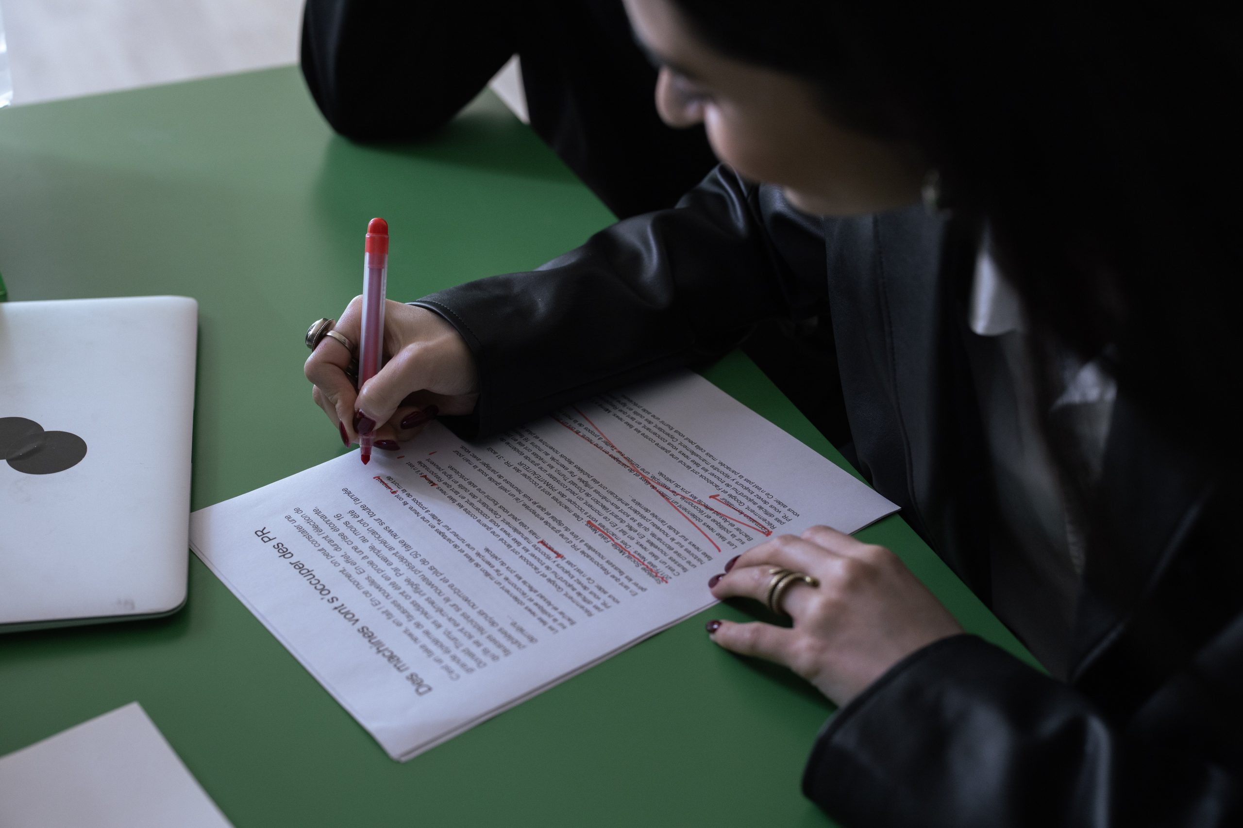 Woman in a dark jacket uses a red pen to edit a document on a green table, illustrating human review of content writing AI tools.